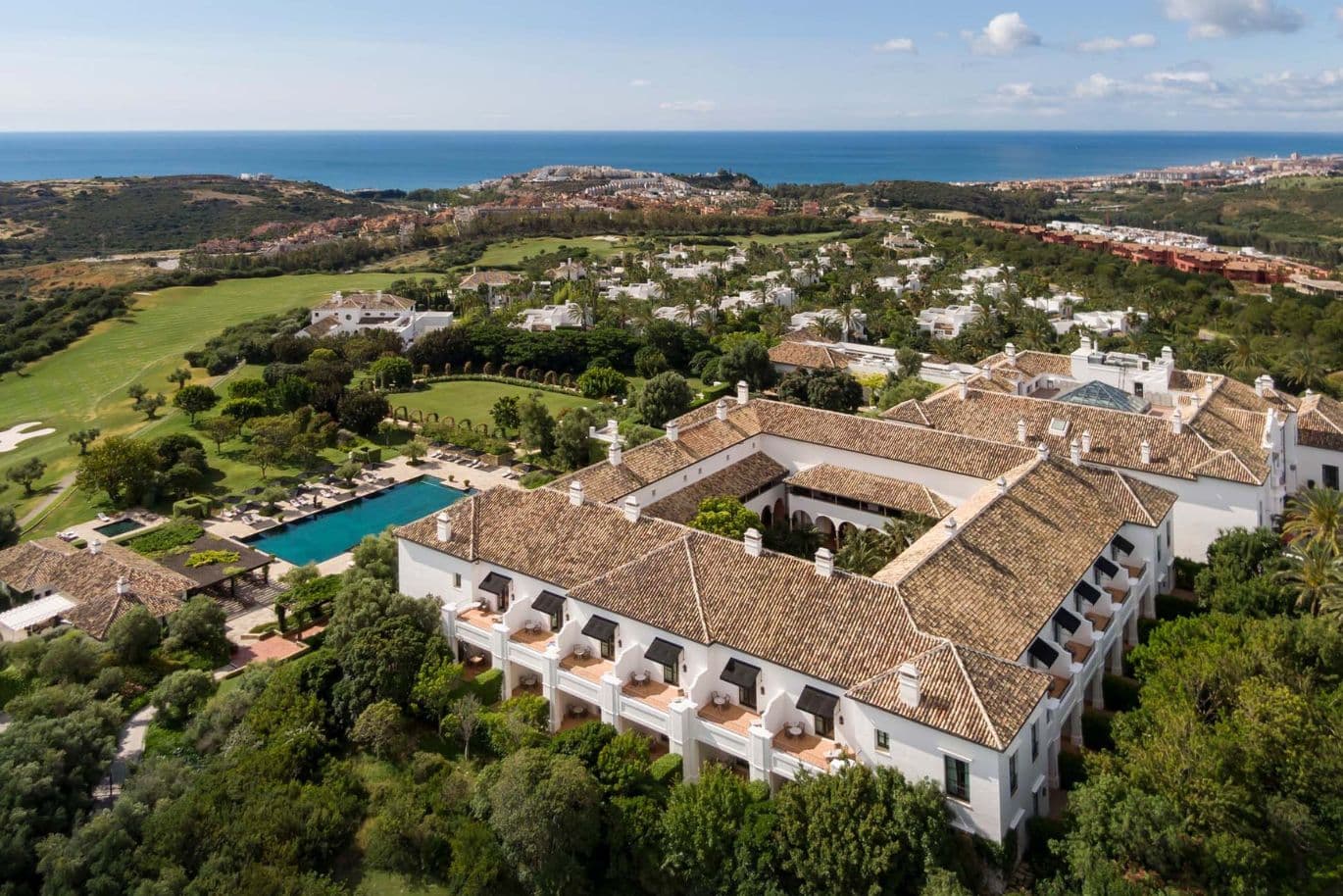 Aerial view of a large, tiled-roof building surrounded by greenery, a golf course, and a pool, with the sea visible in the background.