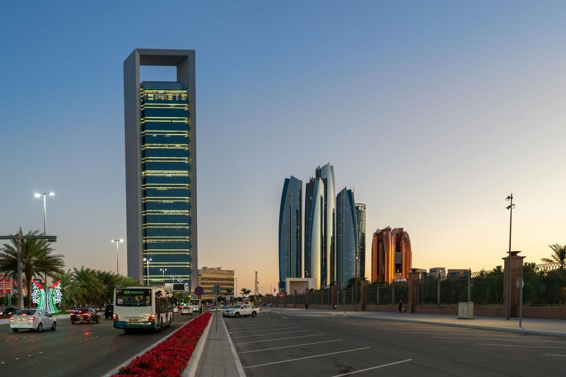 Cityscape at dusk with modern skyscrapers, a lit-up building, palm trees, and traffic on a wide road lined with flowers.