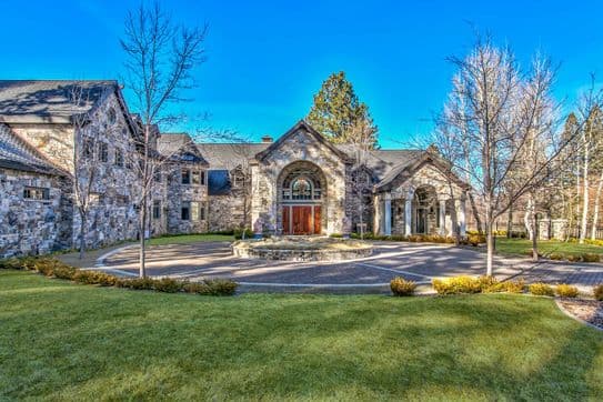 Stone mansion with a grand entrance, arched windows, circular driveway, and manicured lawn under a clear blue sky.