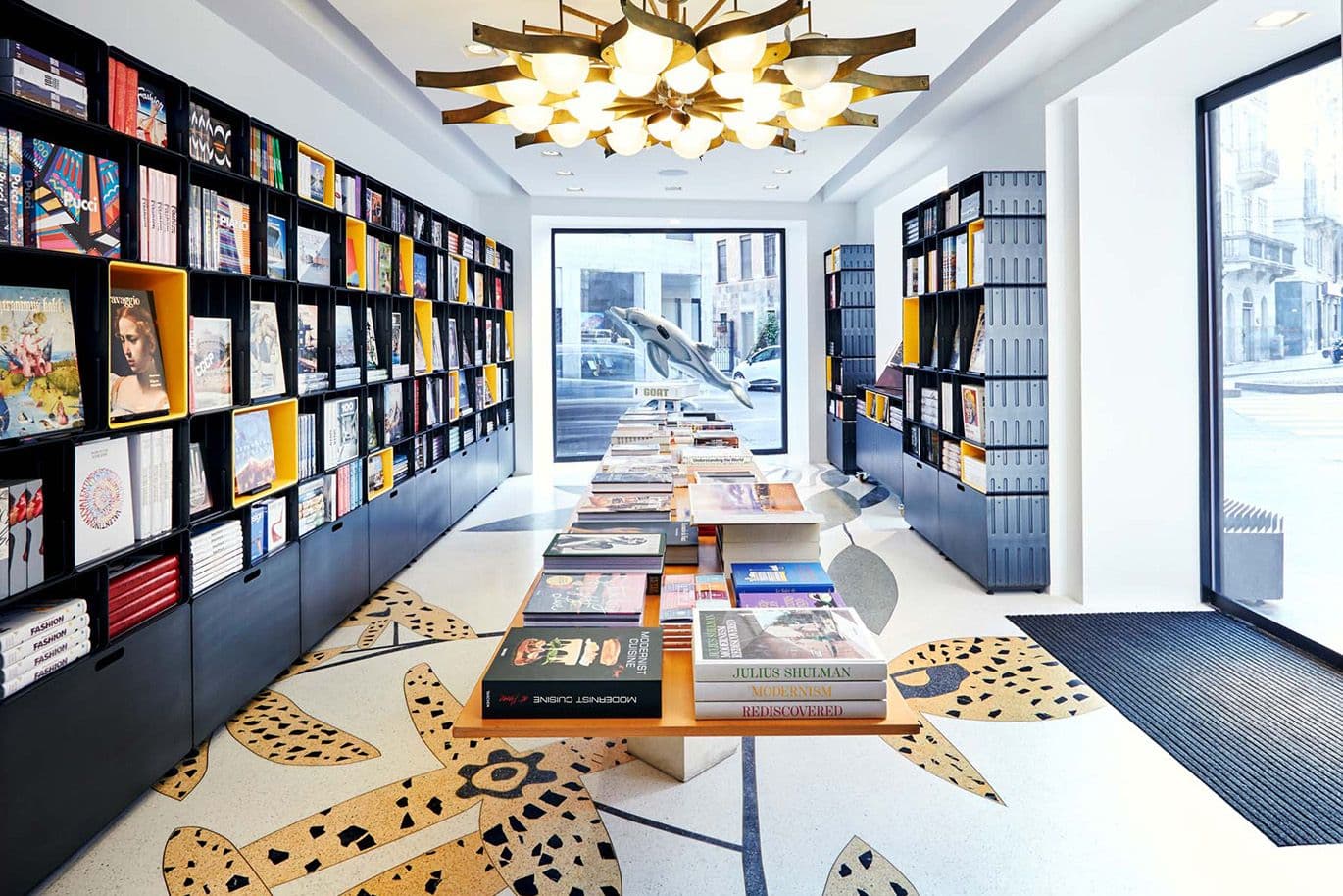 Intérieur moderne d'une librairie avec de longues tables exposant des livres, entourées de hautes étagères. Éclairage au plafond unique et grandes fenêtres.