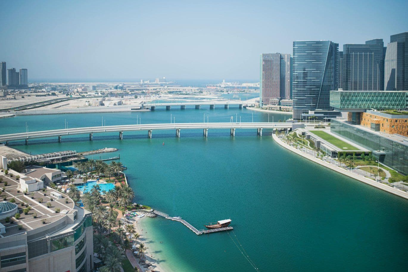Aerial view of Abu Dhabi with modern skyscrapers, a long bridge over a turquoise bay, and palm trees lining the waterfront.