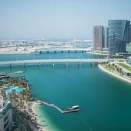 Aerial view of Abu Dhabi with modern skyscrapers, a long bridge over a turquoise bay, and palm trees lining the waterfront.