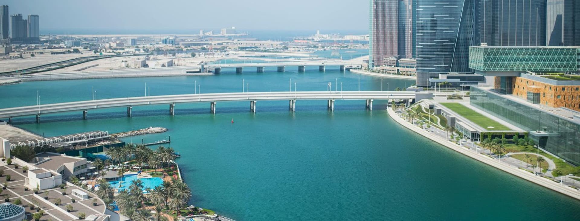 Aerial view of Abu Dhabi with modern skyscrapers, a long bridge over a turquoise bay, and palm trees lining the waterfront.