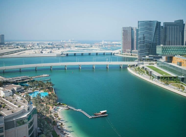 Aerial view of Abu Dhabi with modern skyscrapers, a long bridge over a turquoise bay, and palm trees lining the waterfront.