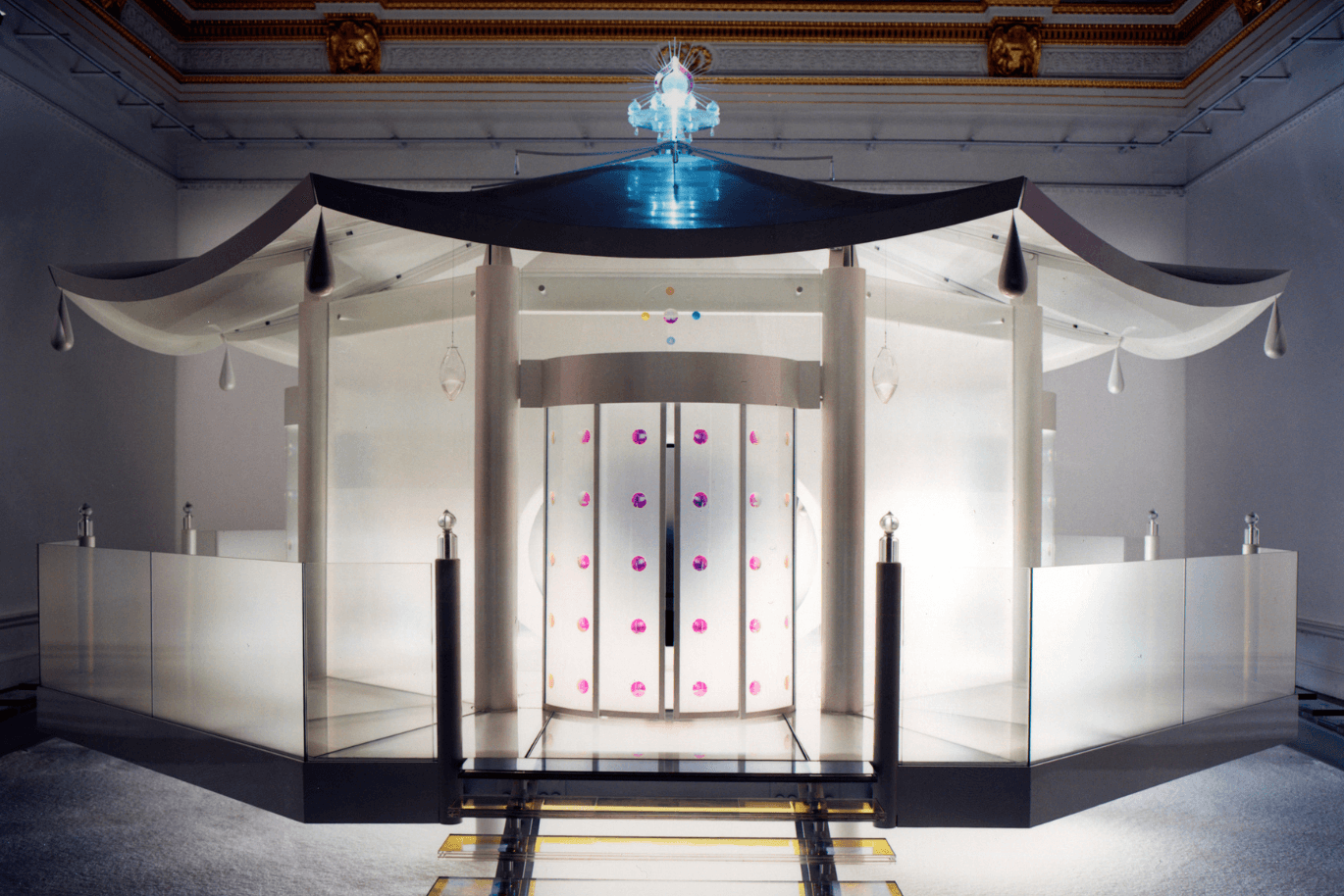 Ein moderner, lichtdurchfluteter Pavillon mit geschwungenem Dach, hängenden Tropfen und farbenfrohen runden Verzierungen, eingebettet in eine elegante Umgebung.