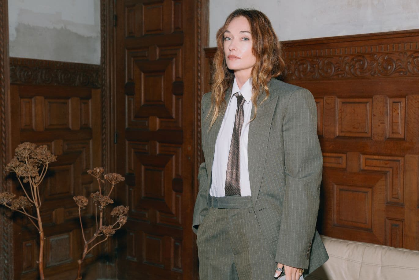 Person in a green plaid suit and tie stands in a vintage room with ornate wooden paneling and dried flowers.