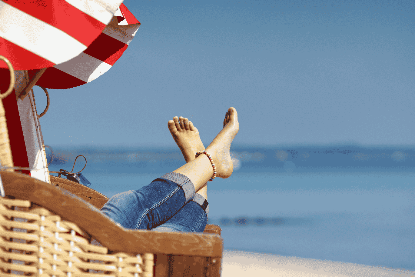 Person relaxing on a beach chair under a red and white umbrella, feet up, wearing jeans, with a view of the ocean in the background.