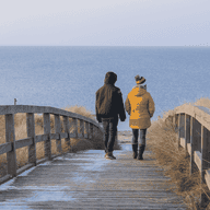 Two people in winter clothing walk on a wooden boardwalk towards the sea, surrounded by dry grass under a clear sky.