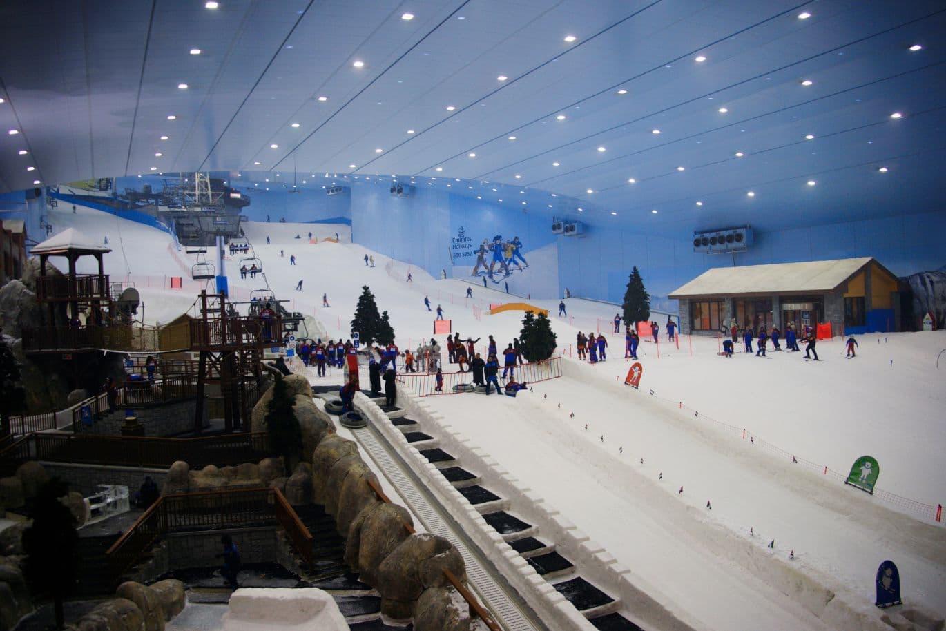 Indoor ski resort with skiers and snowboarders on snowy slopes, a chairlift, wooden structures, and snow play areas inside a large enclosed facility.