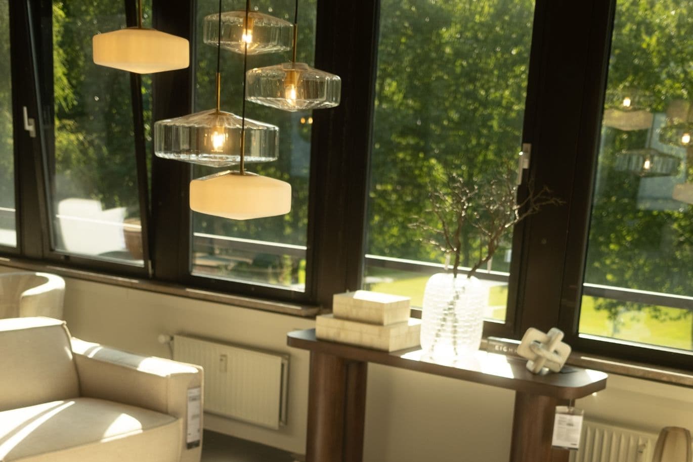 Bright living room with beige sofas, a large ottoman, pendant lights, and a window overlooking greenery. Sunlight streams in, creating a cozy atmosphere.