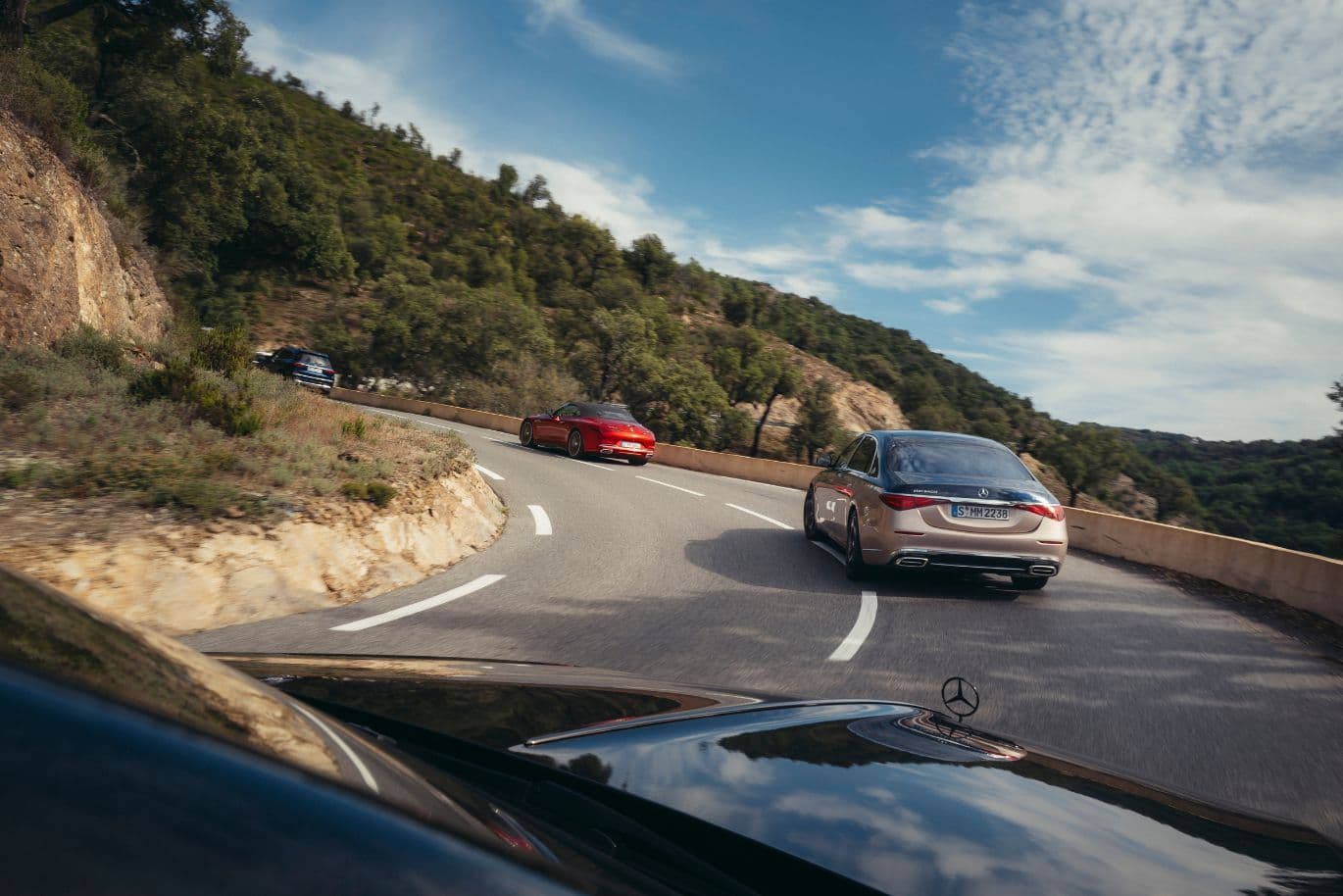 Autos fahren auf einer kurvenreichen Bergstraße mit Bäumen und einem teilweise bewölkten Himmel im Hintergrund.