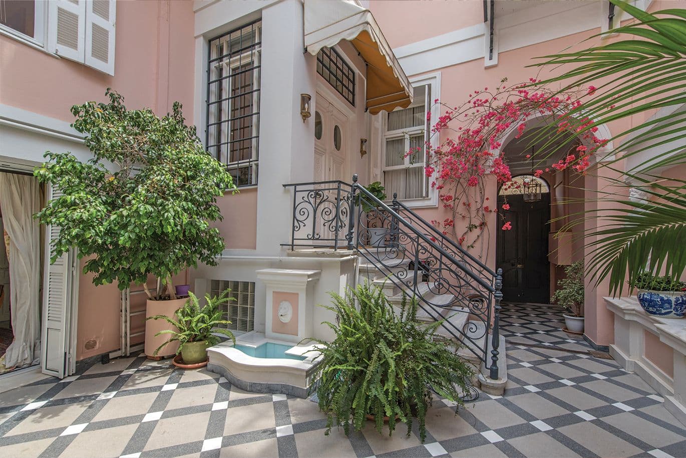 Νεοκλασική αυλή κατοικίας στην Αθήνα με ροζ τοίχους, σιδερένια κιγκλιδώματα και φυτά, χαρακτηριστικό παράδειγμα αθηναϊκής αρχιτεκτονικής.