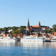 A scenic view of Flensburg's waterfront with boats docked, historic buildings, and a tall church tower under a clear blue sky.