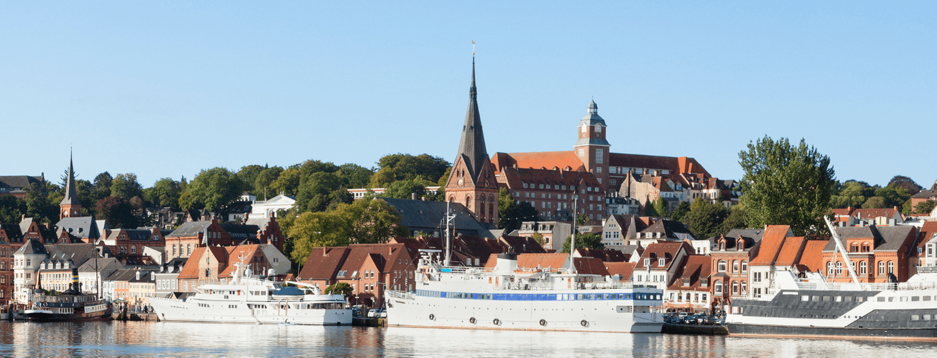 A scenic view of Flensburg's waterfront with boats docked, historic buildings, and a tall church tower under a clear blue sky.