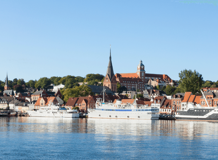A scenic view of Flensburg's waterfront with boats docked, historic buildings, and a tall church tower under a clear blue sky.