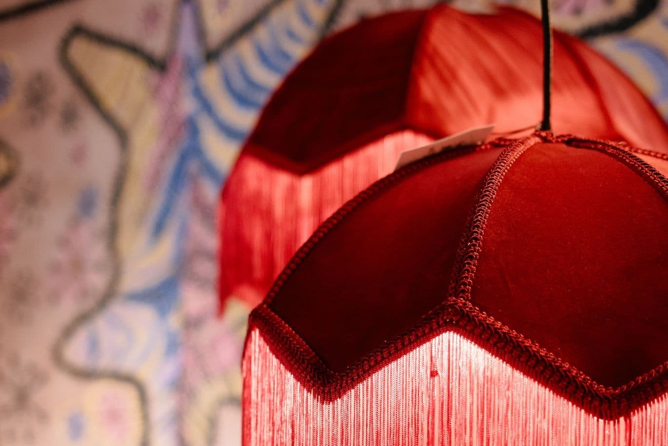 Close-up of two red, fringed lampshades with a hexagonal pattern against a colorful, blurred background.