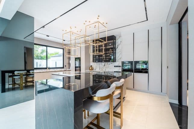 Modern kitchen with a sleek black marble island, white cabinets, and geometric gold lighting fixtures. Large windows offer ample natural light.