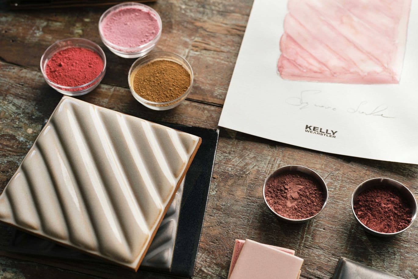 Keramikfliese mit diagonalen Rillen, umgeben von kleinen Schälchen mit Pigmentpulver und einem Aquarellmotiv auf Papier.