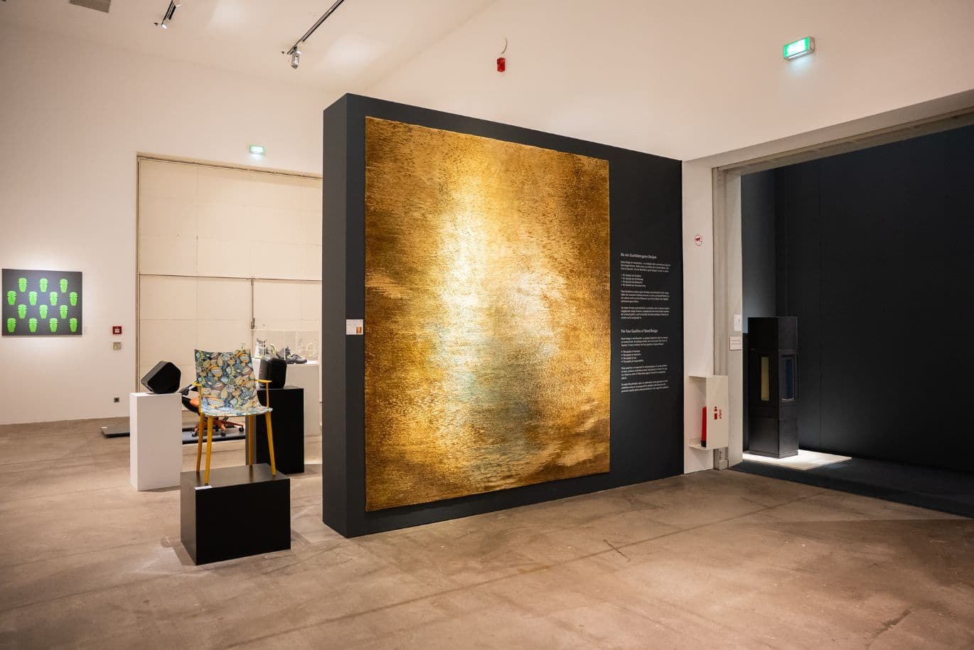 Innenraum einer Kunstgalerie mit großem goldenen abstrakten Gemälde an dunkler Wand; verschiedene moderne Kunstwerke auf Podesten und an Wänden ausgestellt.