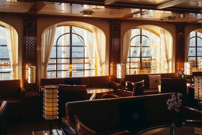Elegante Schiffslounge mit warmer Beleuchtung, weichen Sofas, runden Tischen und großen Fenstern, die einen Blick auf das Meer und die Silhouetten eines Schiffes bieten.