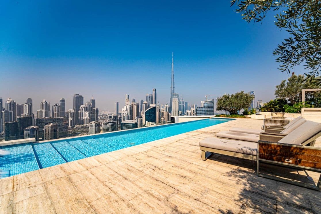 Rooftop infinity pool with city skyline view, featuring tall skyscrapers under a clear blue sky. Sun loungers are placed along the poolside.
