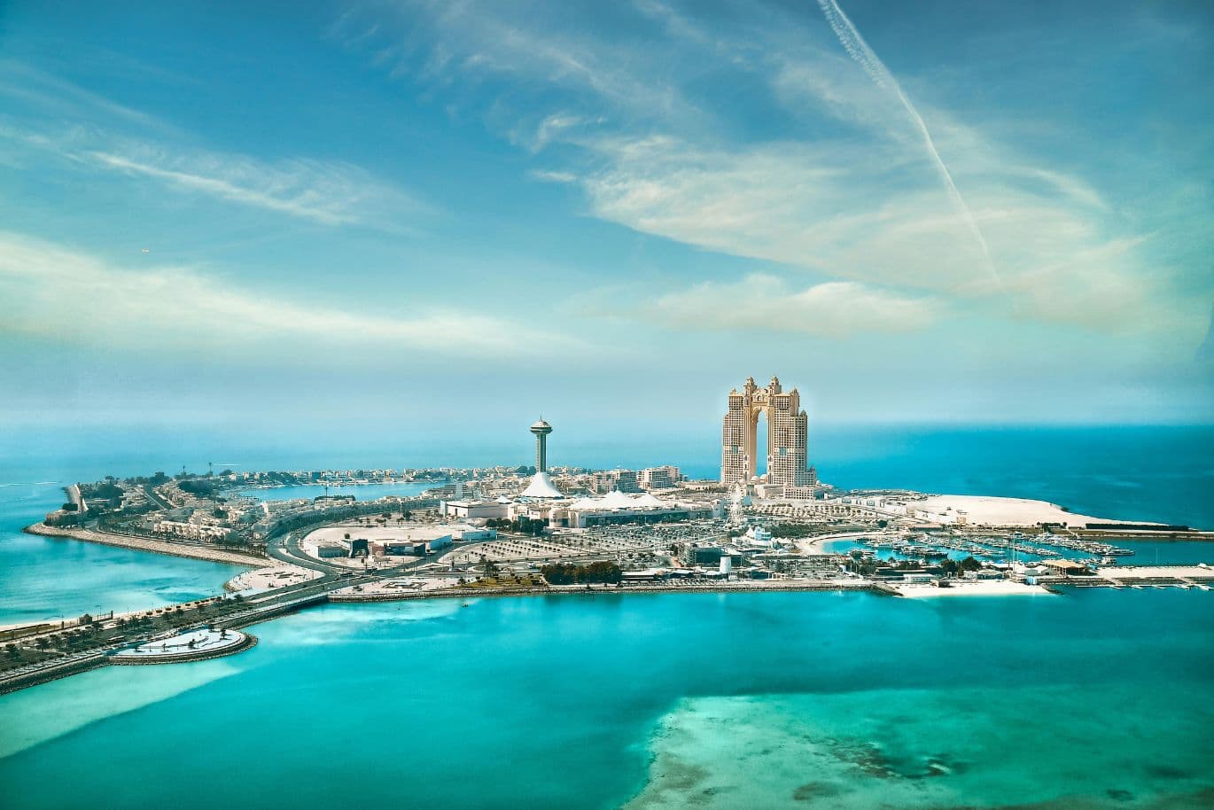 Aerial view of a coastal cityscape with tall buildings, a prominent tower, and a vibrant sea under a clear blue sky.