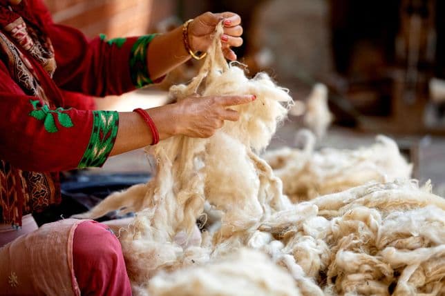 Eine Person in farbenfroher Kleidung verarbeitet Rohwolle, umgeben von Haufen unverarbeiteter Fasern, in einer Textilwerkstatt.