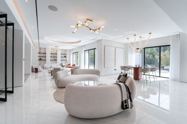 Modern living room with elegant white furniture, round sofas, glass tables, large windows, and sleek lighting. Light marble floors enhance brightness.