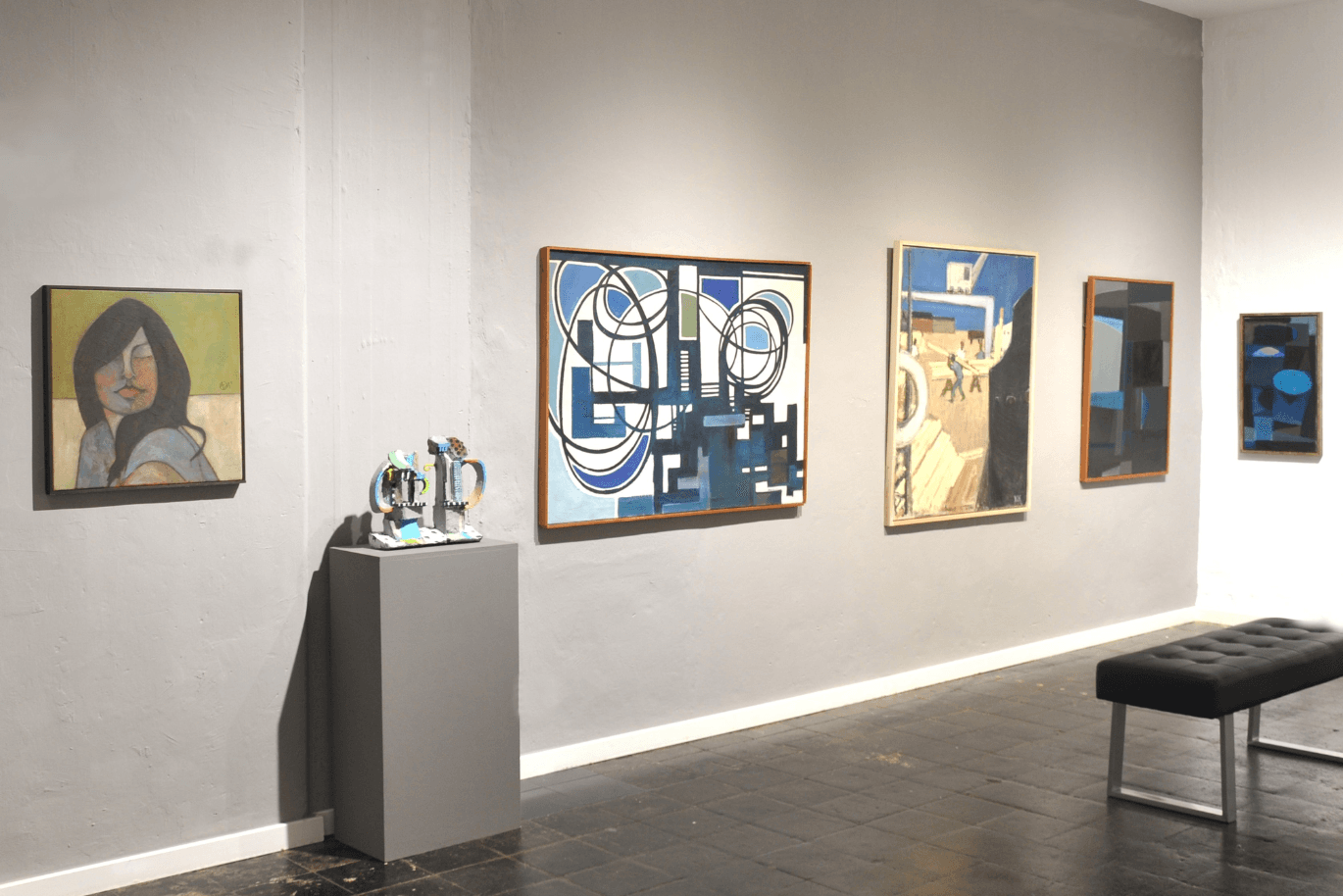 In the Rieck Gallery, pictures with narrow wooden frames hang on a light gray wall. Two sculptures stand on a pedestal.