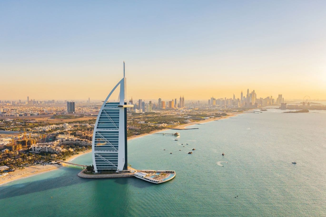 Aerial view of Burj Al Arab on an artificial island, with Dubai's skyline and coastline under a clear sky at sunset.