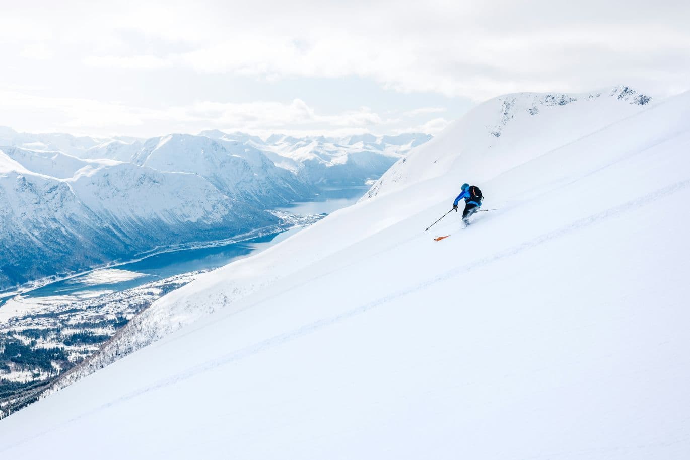 A skier in blue descends a snowy mountain slope with a scenic view of distant snow-covered peaks and a winding river below.