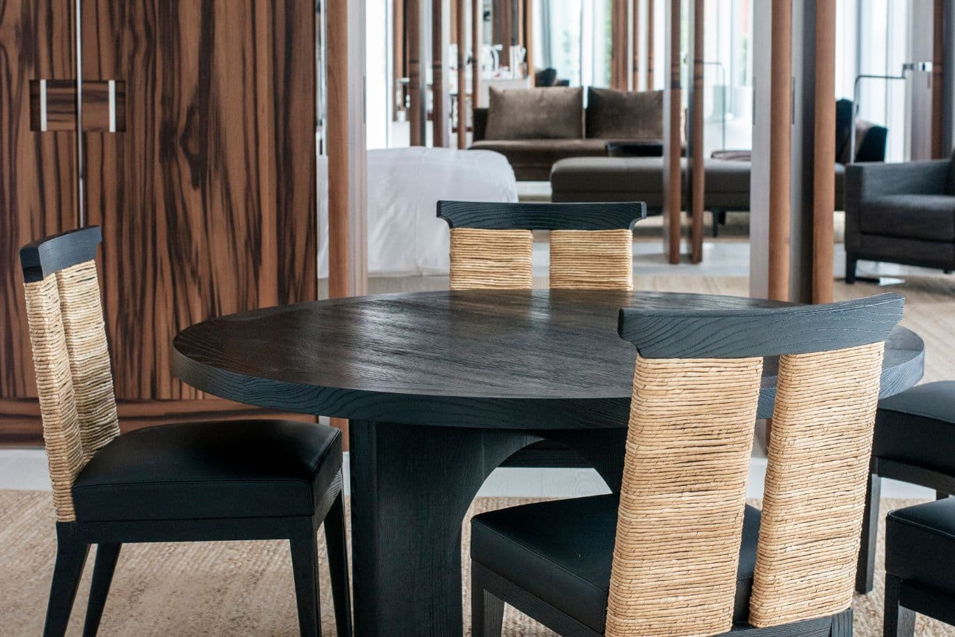 A round wooden dining table with four chairs featuring woven backs, set in a modern room with wooden paneling and a glimpse of a bed.