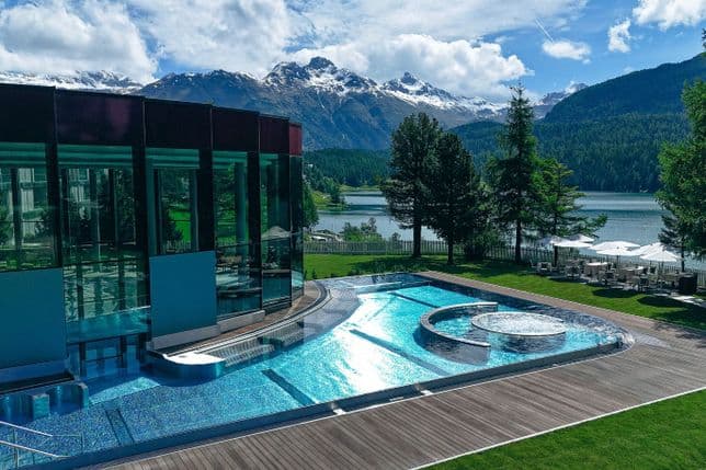 A modern glass building overlooks an outdoor pool with a circular section, set against mountains, a lake, and lush greenery under a blue sky.