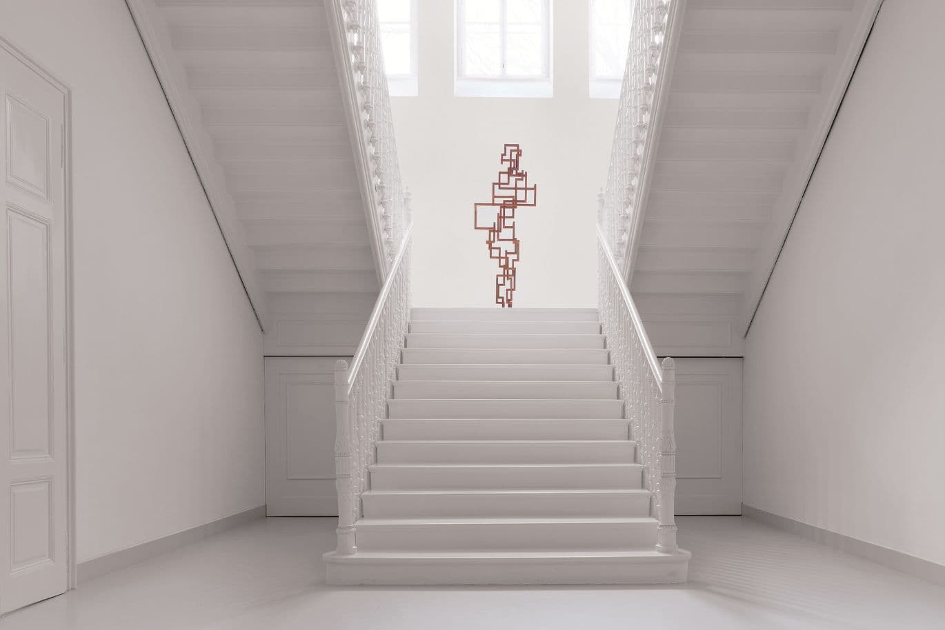 Heller, symmetrische Treppe mit weißen Geländern; vor Fenstern hängt zentral eine rote Wandskulptur.