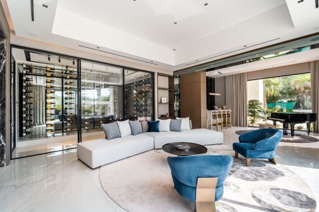 Spacious living room with white sectional sofa, blue armchairs, round coffee table, wine display, and large windows overlooking greenery.
