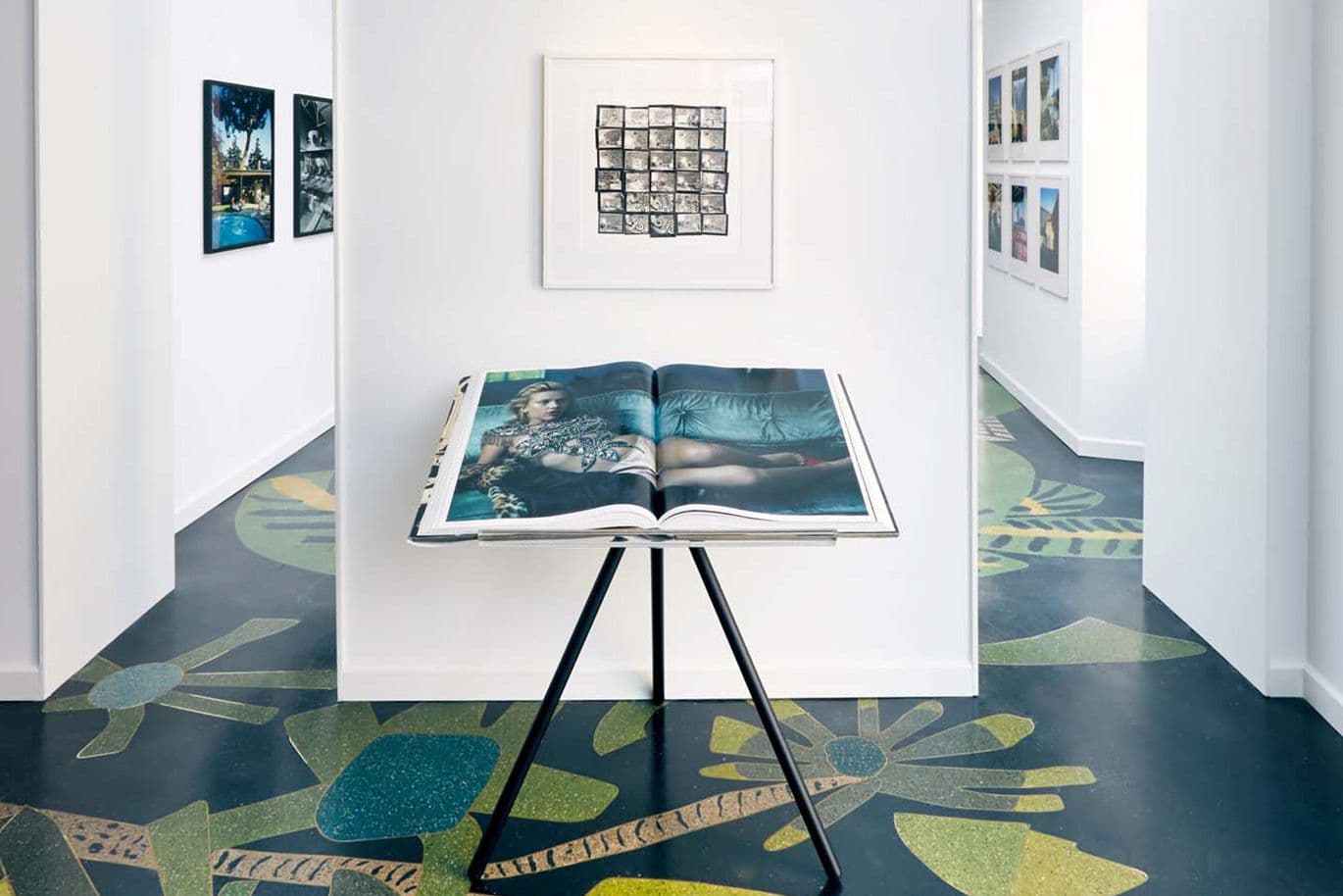 Ouvrez un livre d'art posé sur un trépied dans une galerie où sont exposées des œuvres encadrées sur des murs blancs et un sol coloré à motifs.