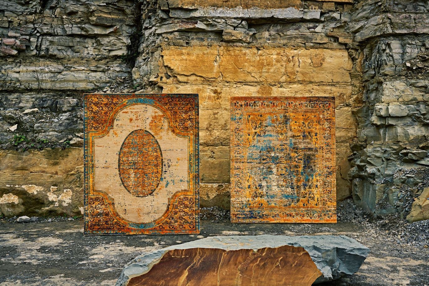 Zwei kunstvoll verzierte Teppiche mit aufwendigen Mustern und gealterter Struktur sind vor einer rauen Steinwand ausgestellt, im Vordergrund befindet sich ein großer Felsbrocken.