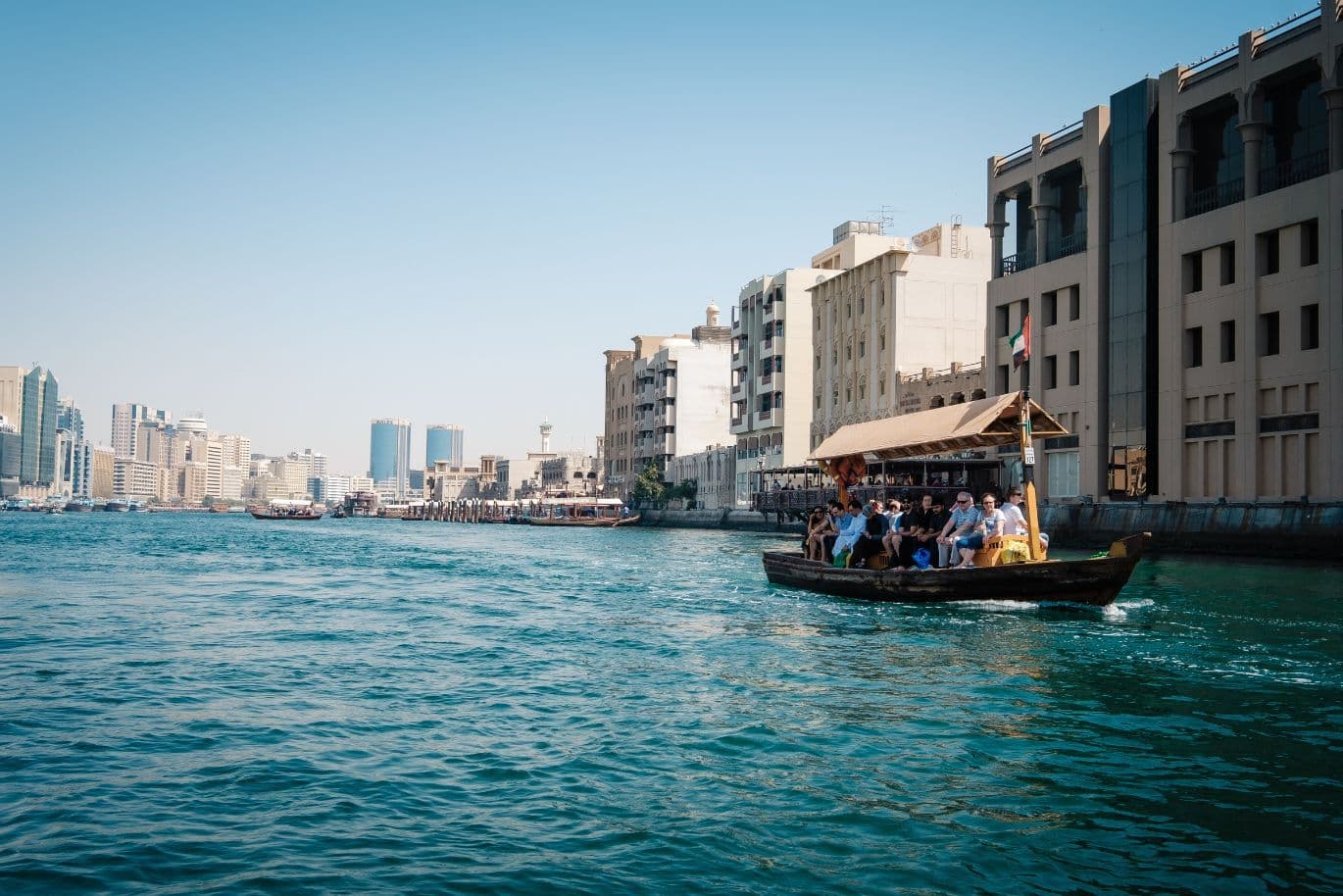 A small boat with passengers sails on Dubai Creek, flanked by modern buildings under a clear sky.