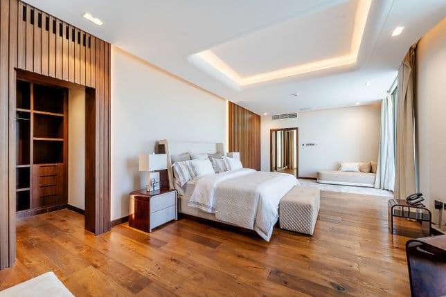 Spacious modern bedroom with wooden floors, neutral tones, a large bed, seating area, and open closet. Bright lighting and a minimalist design.