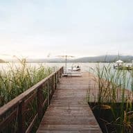 Wooden jetty through reeds on a tranquil lake; at the end, a sun lounger and a white parasol, next to it a moored motorboat. In the background, hills, the shoreline and another jetty in the soft evening light.