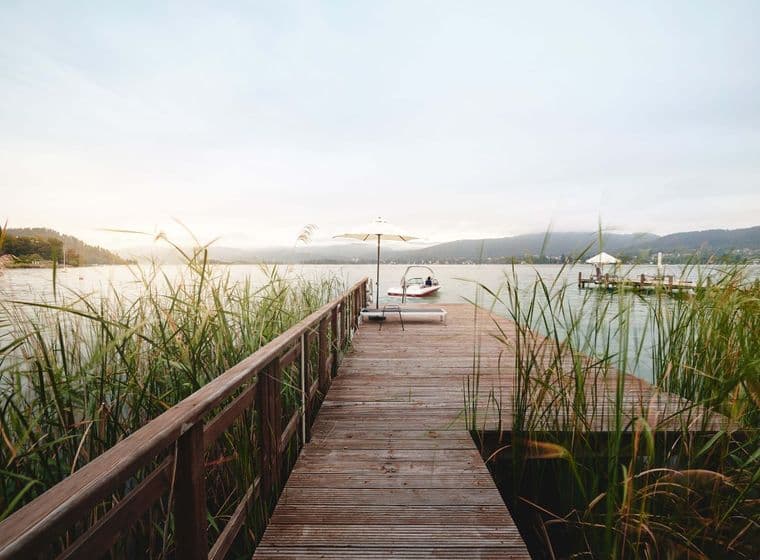Wooden jetty through reeds on a tranquil lake; at the end, a sun lounger and a white parasol, next to it a moored motorboat. In the background, hills, the shoreline and another jetty in the soft evening light.