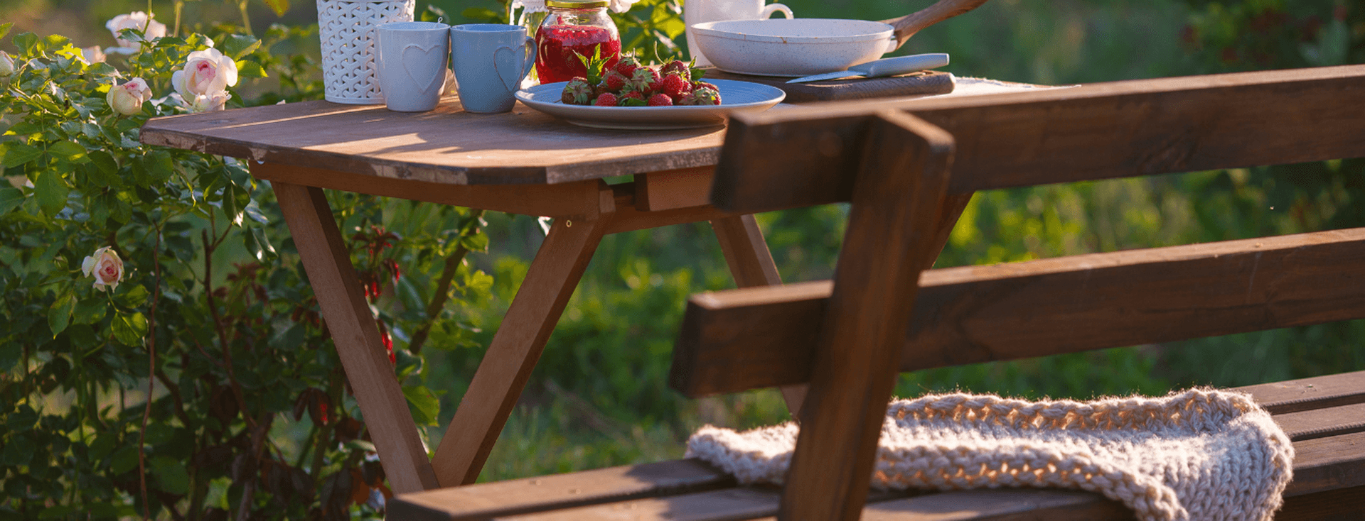 A wooden table with flowers, strawberries, cups, and a plate in a garden setting, next to a bench with a knitted blanket draped over it.
