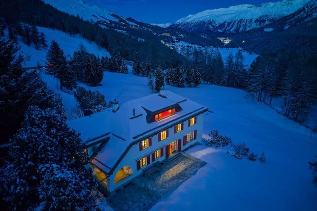 Luftaufnahme eines schneebedeckten Chalets mit warm leuchtenden Fenstern in einem blauen Bergtal in der Dämmerung.