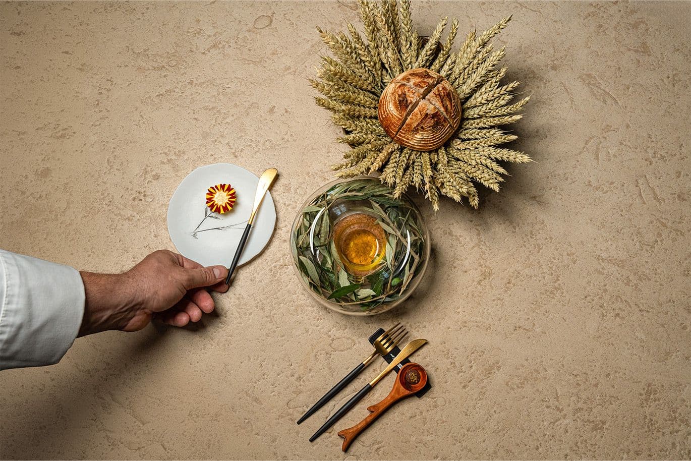 A flat-lay composition showing bread, olive oil, and a small plated bite presented with elegant cutlery on a stone-textured surface.