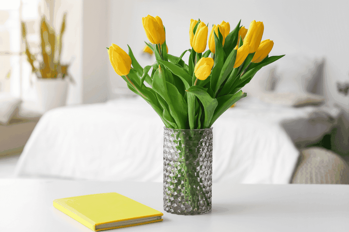 Yellow tulips on a table, a yellow book next to it, a white bed in the blurred background