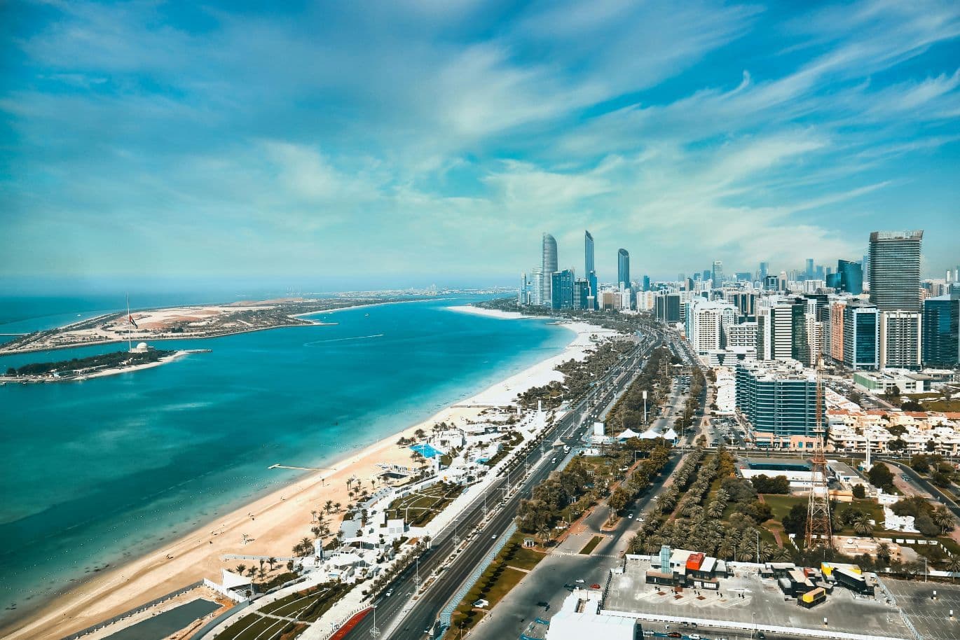 Aerial view of a coastal cityscape with skyscrapers along a sandy beach, set against vivid blue water and a sky with wispy clouds.