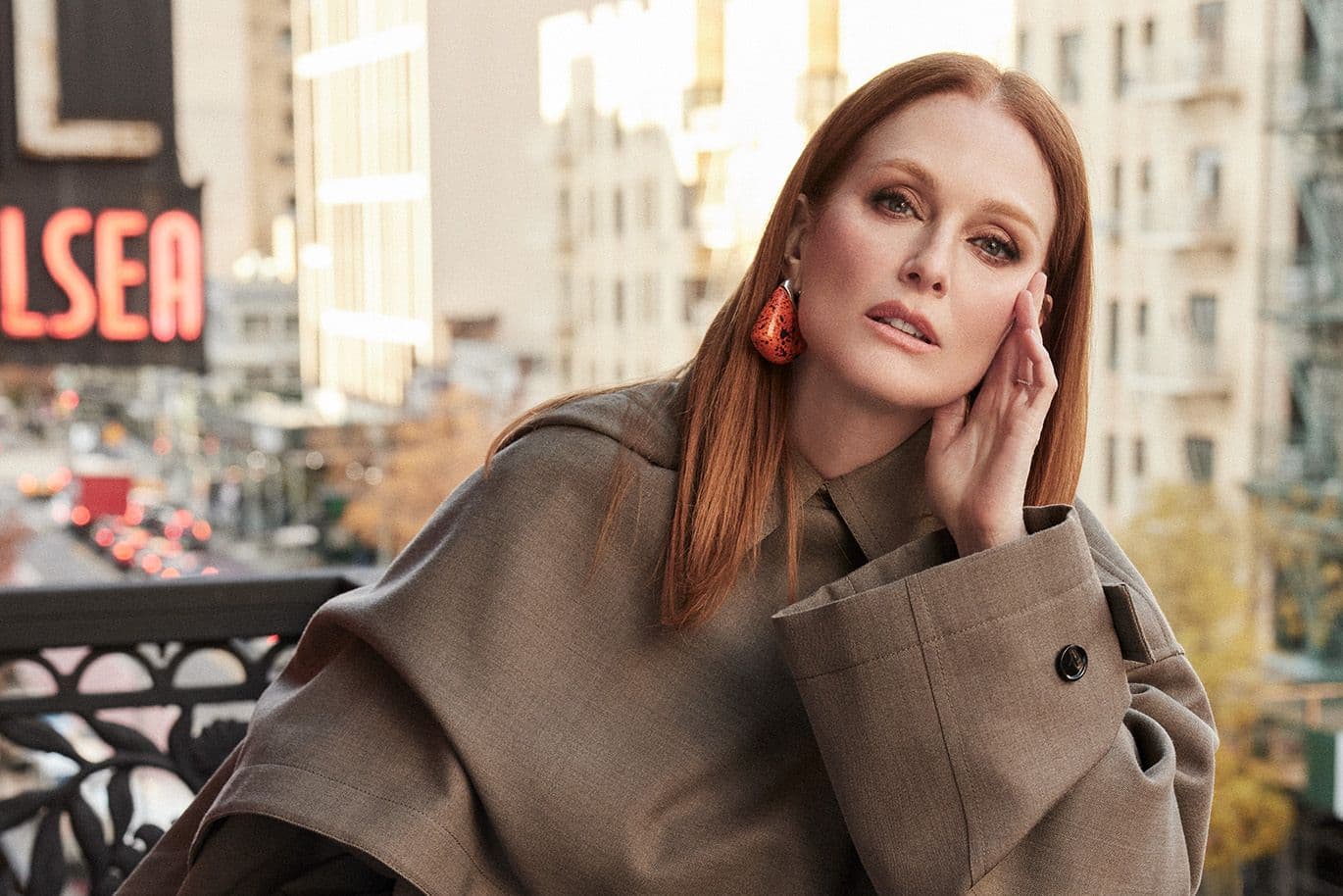 Red-haired woman in a brown coat and orange earrings, leaning on her hand on a city balcony.