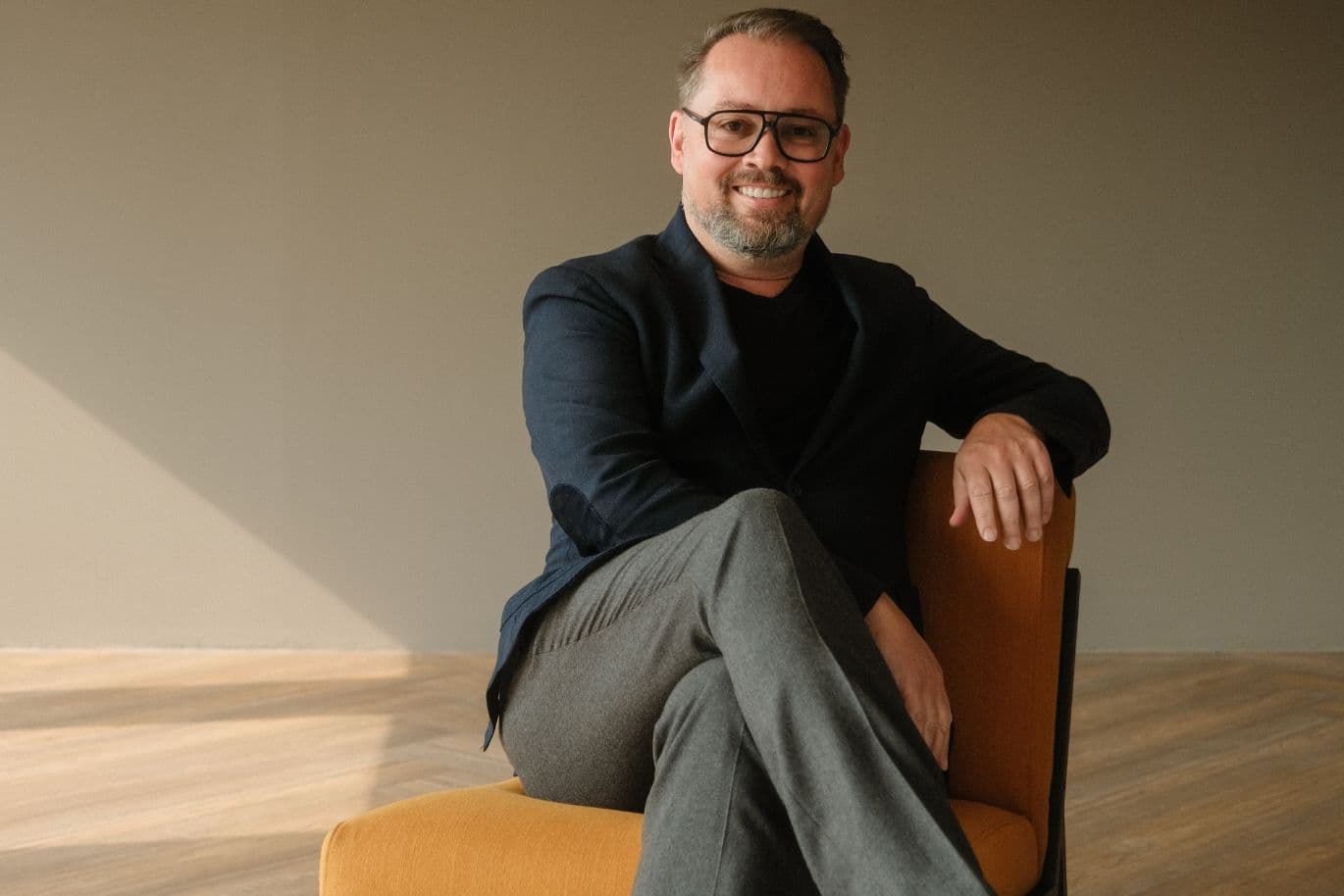 Smiling man with glasses and a beard, sitting on an orange chair and wearing a dark blazer and gray pants, in a warmly lit room.