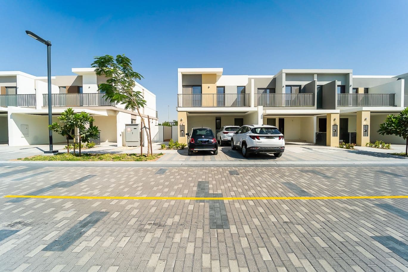 Modern suburban homes with balconies, two cars parked in driveways, and a clear blue sky above.