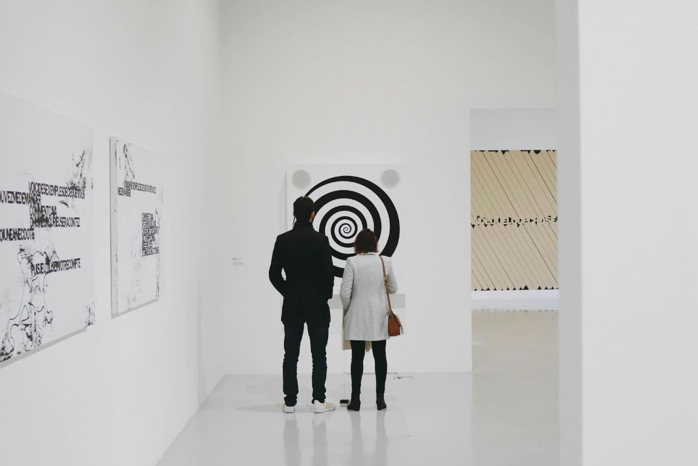 Two people stand in a modern art gallery, observing a black-and-white spiral artwork on a white wall.